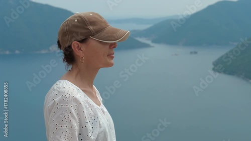 Mature woman enjoying a beautiful fjord landscape view. Happy mature woman in a baseball cap enjoying the serene view of a beautiful fjord, admiring the majestic mountains and tranquil water