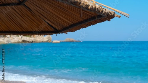 Straw beach umbrella and turquoise ocean waves. Peaceful view from under a straw beach umbrella looking out at the calm turquoise sea with gentle waves washing ashore on a beautiful sunny day
