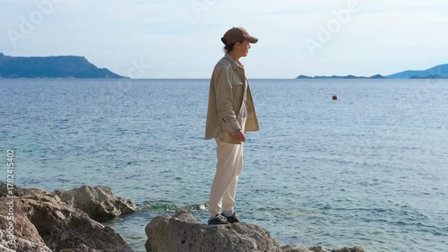 Woman walking on rocks by the sea. Casual-dressed woman wearing a cap carefully balancing and walking across rocky coastal terrain, savoring scenic ocean views during sunny summer day
