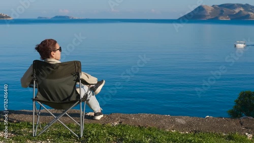 Relax in a camp chair by the sea. Tourist enjoying a peaceful moment, sitting on a camping chair by the sea, observing a distant boat sailing on the tranquil waters with islands