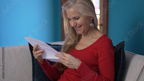 A middle age woman with long blonde hair is sitting on a couch, smiling and holding a letter with good news. Giving a thumbs up, he is wearing a red shirt