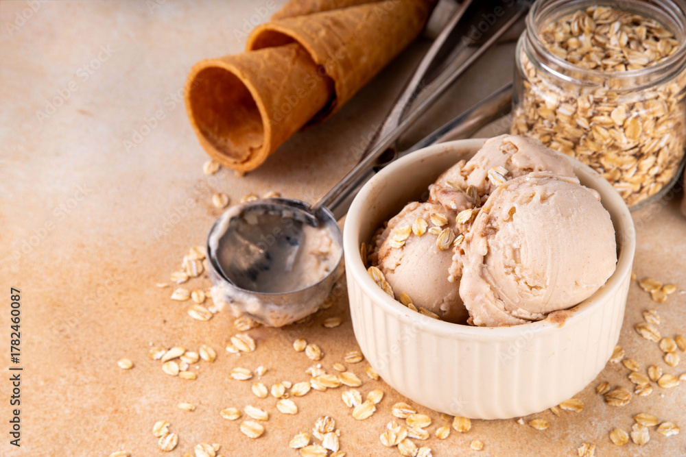 Fototapeta premium Vegan oats milk non-dairy ice cream, scoops of vanilla creamy oatmeal milk gelato in beige bowl, with ice cream waffle cones and rolled oats flakes