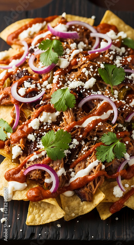Overhead shot of loaded nachos with pulled pork, red onions, cilantro, cheese, and sauce, served on a dark surface.