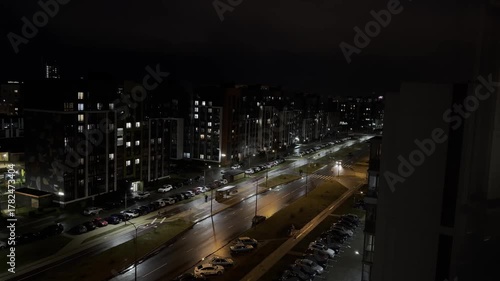 Street at night with illuminated residential buildings, wet asphalt and reflections, cars parked