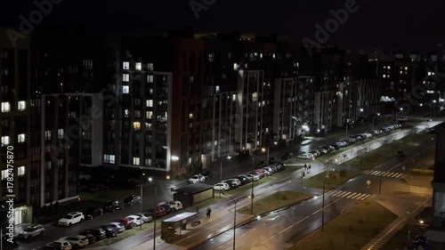 Street at night with pedestrians walking along illuminated buildings, cars parked and moving on wet