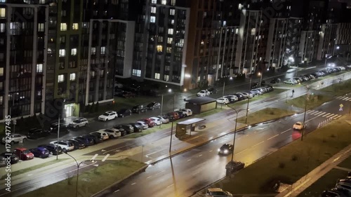 Street with cars and illuminated buildings at night, wet asphalt with reflection, residential