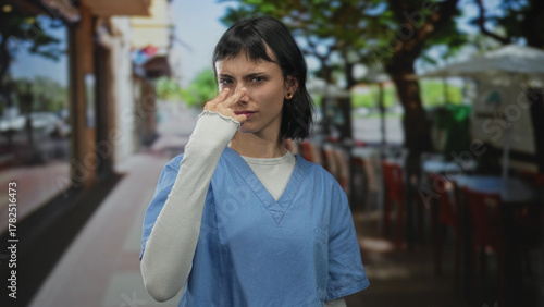 Photography Woman nurse in blue scrubs making a cut throat gesture on a sunlit street sidewalk beside cafe seating and leafy trees; intimidation