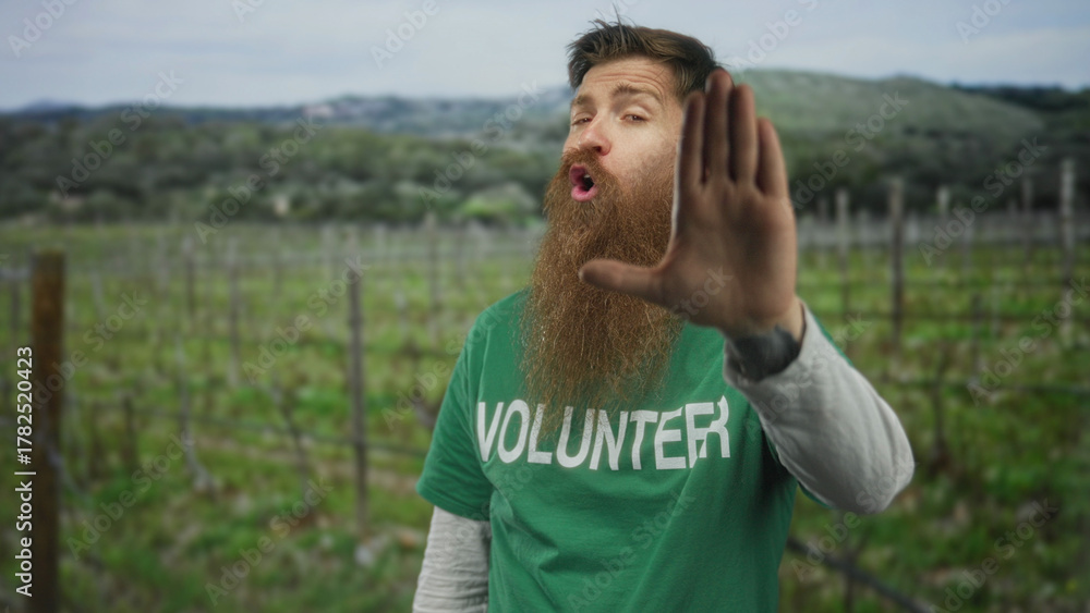 Fototapeta premium Man wearing green volunteer shirt raises hand in stop gesture in a countryside field; safety awareness.