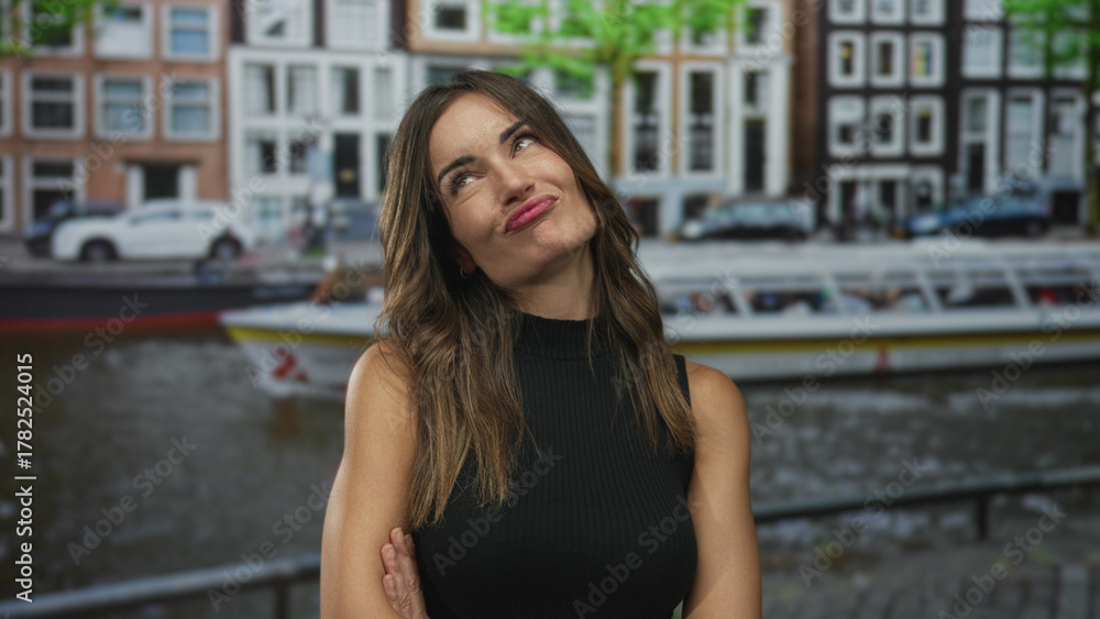 Fototapeta premium Woman with arms crossed leaning on a railing by a canal on a cobbled street among historic buildings in bright daylight; serenity.