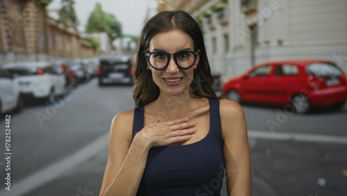 Fotografie Woman wearing glasses and navy tank top clutching chest in apparent heartattack on urban street lined with parked cars and buildings; surprise