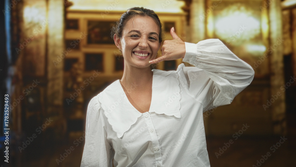 Obraz premium Woman smiling while making a call gesture with hand to ear inside ornate church building with chandeliers and paintings; playful invitation.