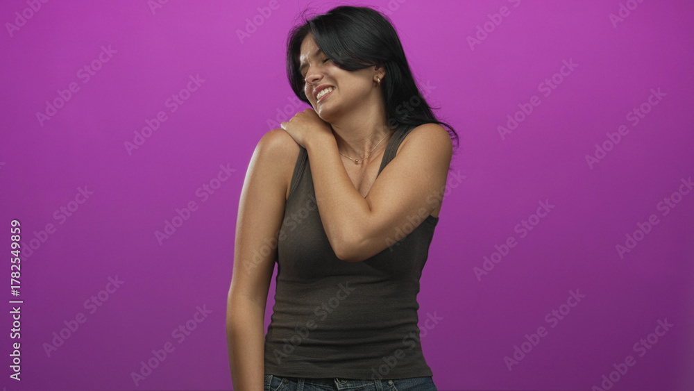 Obraz premium Young hispanic woman in tank top grimacing while holding shoulder in purple studio; muscle strain discomfort.