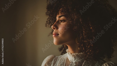 Young woman with voluminous curly hair gazes thoughtfully toward warm side lighting.
