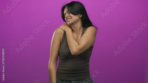 Young hispanic woman in tank top grimacing while holding shoulder in purple studio; muscle strain discomfort.