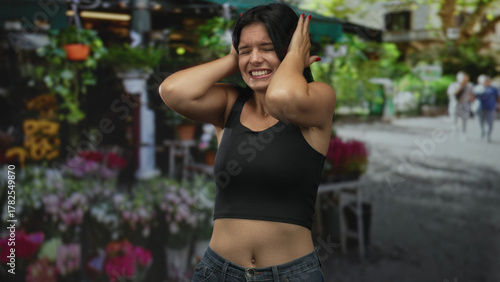 Woman covers her ears with her hands in front of a street flower stall under sunlight while standing near potted blooms; frustration.