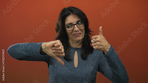 Quadro em tela Middle aged hispanic woman smiling and showing thumbs up and thumbs down in studio with orange backdrop; ambivalence