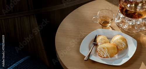 A dish of madeleines with fork and clear glasses jar of herbal’s tea set on wooden table decorated by flowers blow and a book beside the leaning area with pillow in background