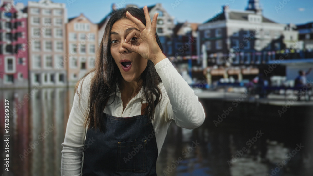 Fototapeta premium Woman makes ok sign around eye while wearing denim apron on amsterdam canal street with historic buildings and water reflections; playful exploration.