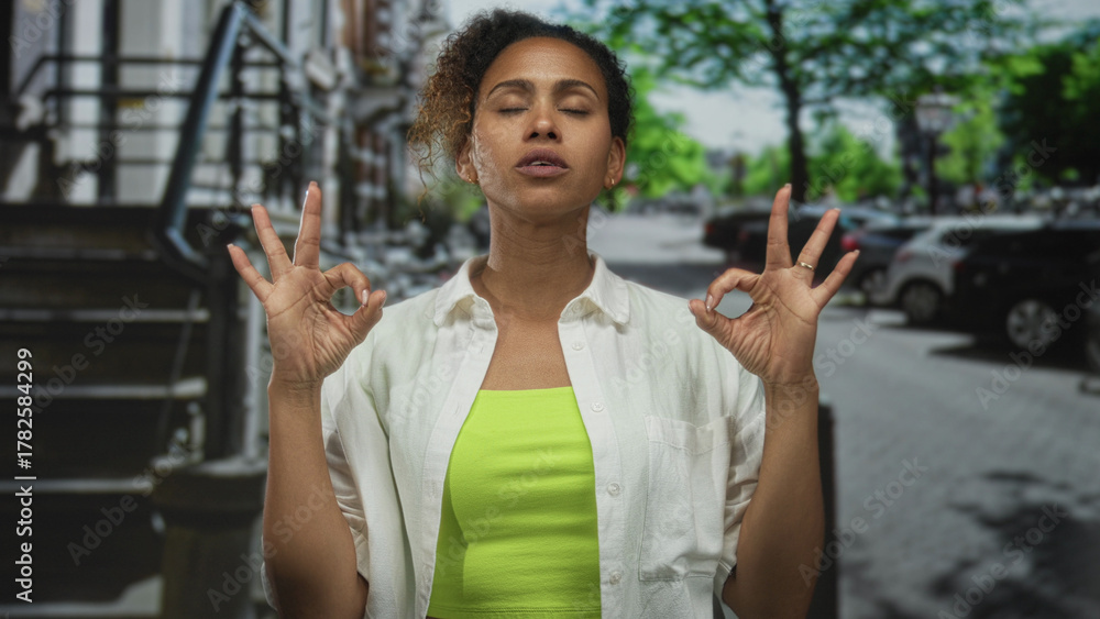 Naklejka premium Woman with closed eyes and raised hands wearing neon green top and white shirt on a street lined with parked cars; calm concentration balance.