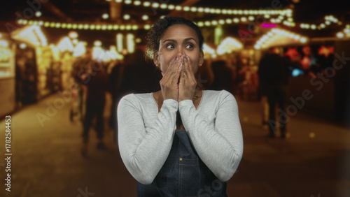 Woman covering mouth with hands in a street market stall aisle under warm string lights; surprise holiday shopping.