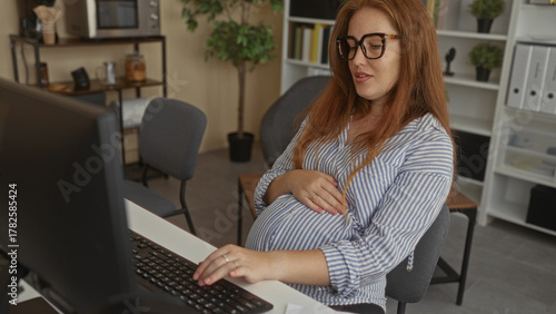 Photos Pregnant woman wearing glasses holds belly at building desk while typing on computer in an office workspace; anticipation
