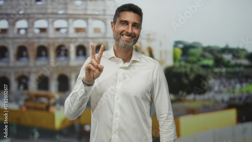 Canvas Print Man smiling and holding up two fingers shows peace sign with right hand in front of building coliseum outdoors, white shirt visible; peace travel