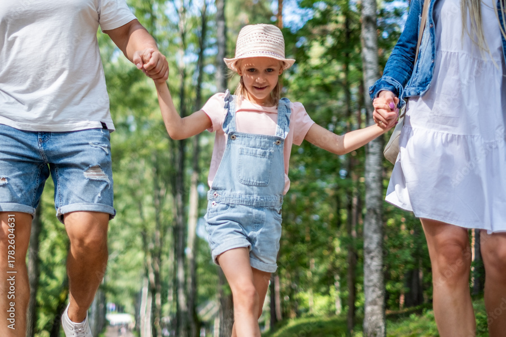 Fototapeta premium Father, mother and daughter walking hand in hand through a lush green forest, enjoying playful moments together, celebrating family bonds and childhood happiness