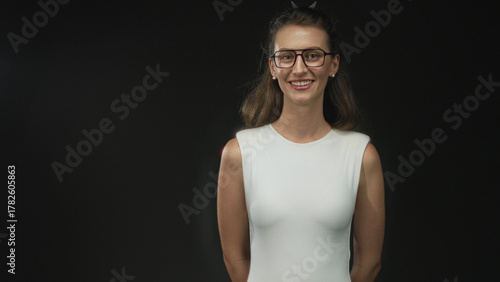 Wallpaper Mural Woman smiling with bare shoulder and hands behind back wearing white sleeveless top and glasses in black studio; joy warmth openness. Torontodigital.ca