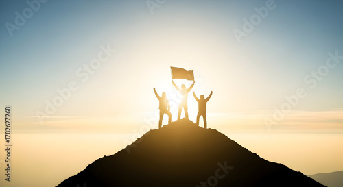 Silhouette of three people celebrating on a mountain top with a flag at sunset or sunrise