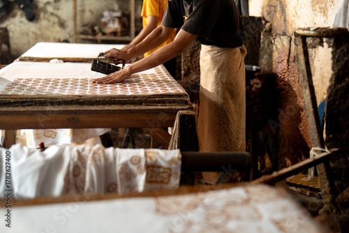 Indonesian batik workers hand stamping wax pattern on white fabric inside traditional workshop, showing teamwork and heritage textile process.