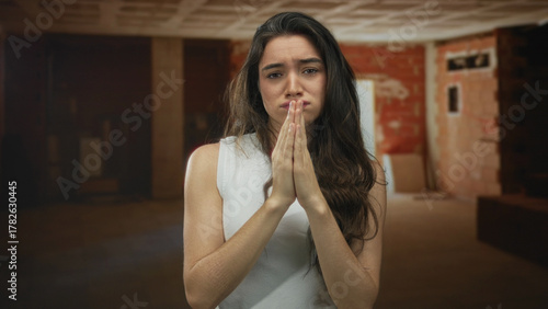 Fotografie Young hispanic woman pressing hands in prayer at construction site interior; desperate plea for relief