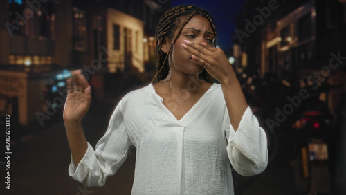 Photos African american woman pinches nose with hand, wincing on dim city street at night; disgust discomfort