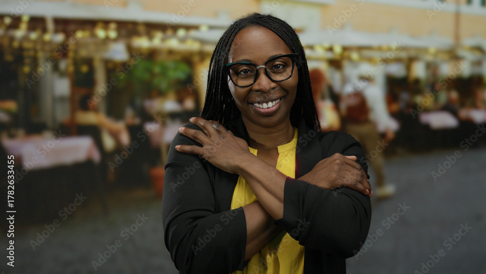 Fototapeta premium Woman smiling with arms crossed in a bustling outdoor market background, blending urban vibes with a warm expression, wearing glasses and a casual outfit