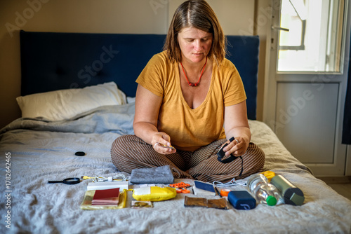 Middle-aged woman checking headlamp operation while assembling emergency go-bag for dangerous emergency situation on bed in bedroom, real moments