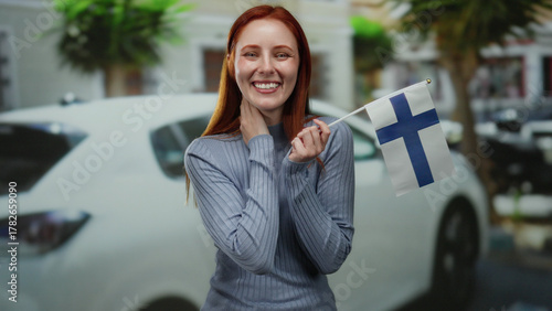Canvastavla Woman smiling outdoors holding finnish flag in city street with red hair and wearing blue sweater, radiating joy and national pride against urban backdrop