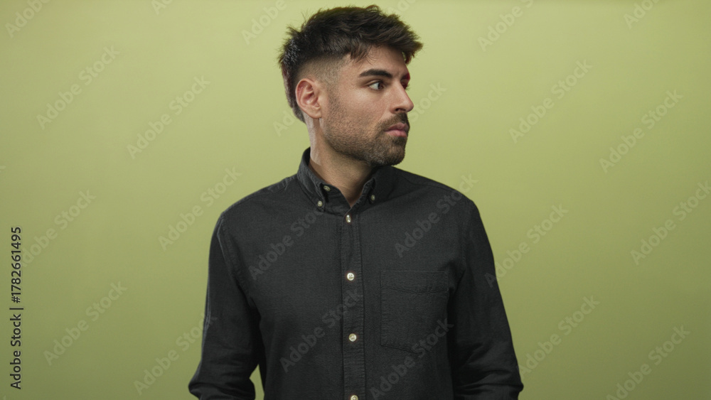 Obraz premium Young hesitant hispanic man buttoning shirt collar with both hands against light green wall in studio; fear anxiety dread worry.