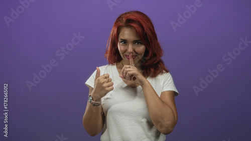 Photos Young hispanic woman in white tee showing thumbs up and finger to lips in purple studio, wearing watch and layered necklaces; playful secret approval