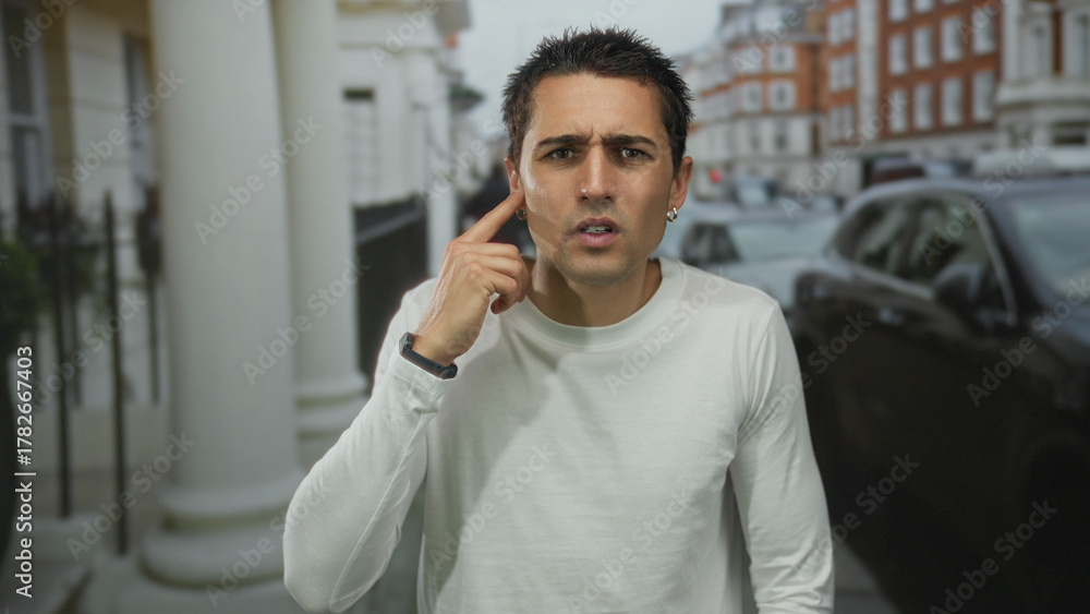 Fototapeta premium Young hispanic man in white long sleeve shirt holds finger to ear on bustling urban street with parked cars; curiosity.