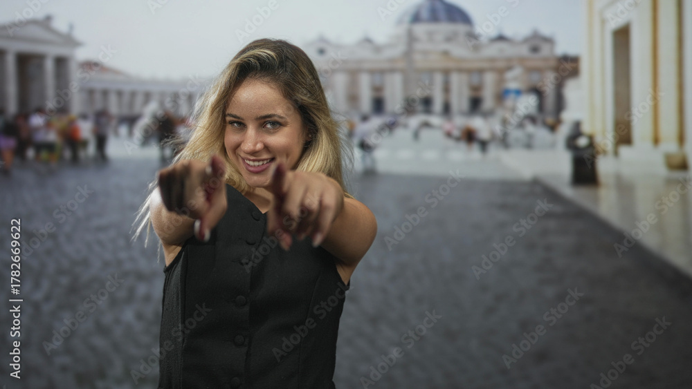Obraz premium Woman points finger at camera with both hands in square at the vatican, smiling and leaning forward; playful confidence.