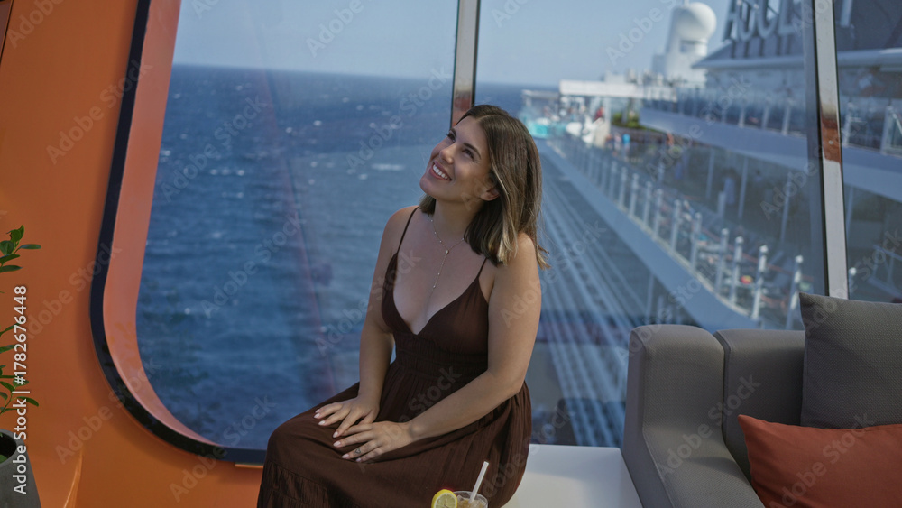 Naklejka premium Woman brunette young hispanic sitting with hands on lap and visible cleavage wearing brown dress by cruise terrace window with ocean view; serenity relaxation.
