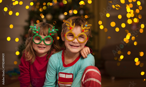 Two kids brother and sister Wearing carnival glasses with Christmas decor on near the Christmas tree at home. 