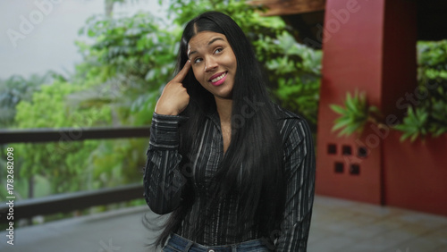 Latin woman in hotel building gestures with finger to head while standing indoors with a playful grimace; craziness.