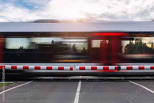Фотография Fast modern regional commute train rushes past closed railway crossing under cloudy sky  sunlight sunset evening