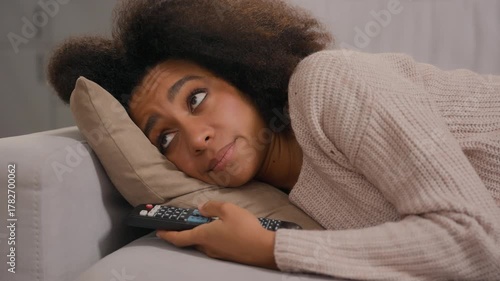 African American tired woman feeling bad negative mood girl lying on sofa living room home holding remote controler switch channels watching television TV at home frustrated emotion exhausted female