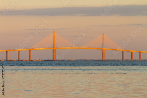 The Sunshine Skyway Bridge stretches gracefully across Tampa Bay at dawn, bathed in soft golden light as the new day begins. Gentle ripples on the calm water mirror the vibrant hues of the morning sky