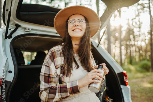 Young caucasian woman sitting in the car trunk in the forest drinking water after a hike during golden hour