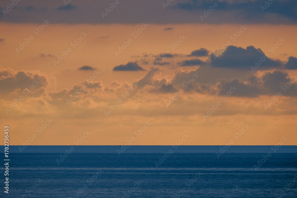 Obraz premium A serene seascape captures a warm sunset spilling pink and orange hues across the sky while the blue ocean stretches to the horizon, inviting calm, reflection, and quiet contemplation.