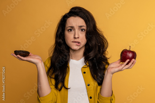 Schilderij op canvas Confused dark-haired cute lady choosing between a high-calorie cookie and a low-calorie fruit during the studio photo shoot