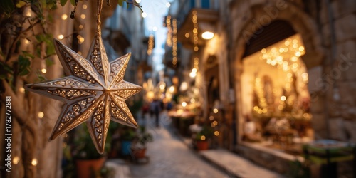 Fototapeta Naklejka Na Ścianę i Meble -  Bethlehem star decoration hangs on a lit street in a southern European town with stone buildings and holiday lights. Christmas atmosphere in a warm historic setting with festive decor.