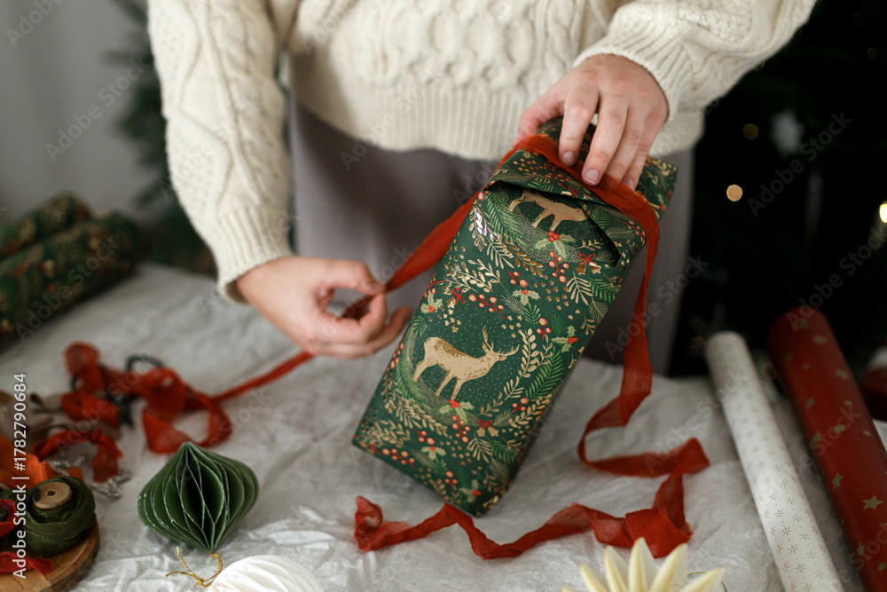 Fototapeta premium Woman in cozy sweater wrapping christmas presents on rustic table with festive paper, modern ornaments, ribbons and scissors against decorated christmas tree. Merry Christmas!
